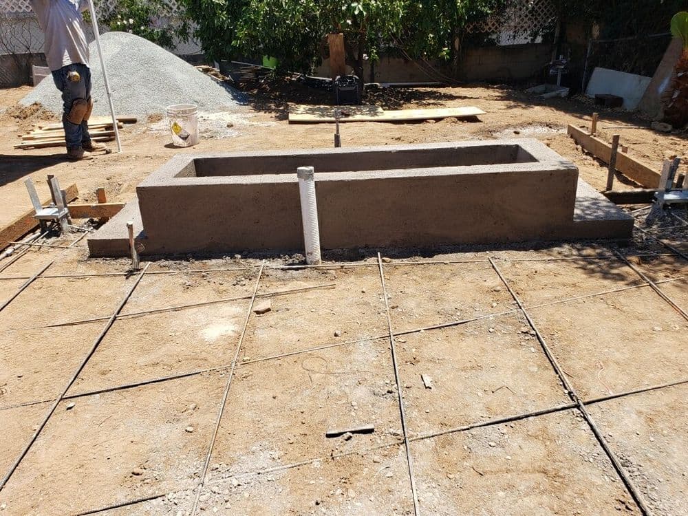 Concrete foundation under construction with reinforcement bars, surrounded by gravel and tools.