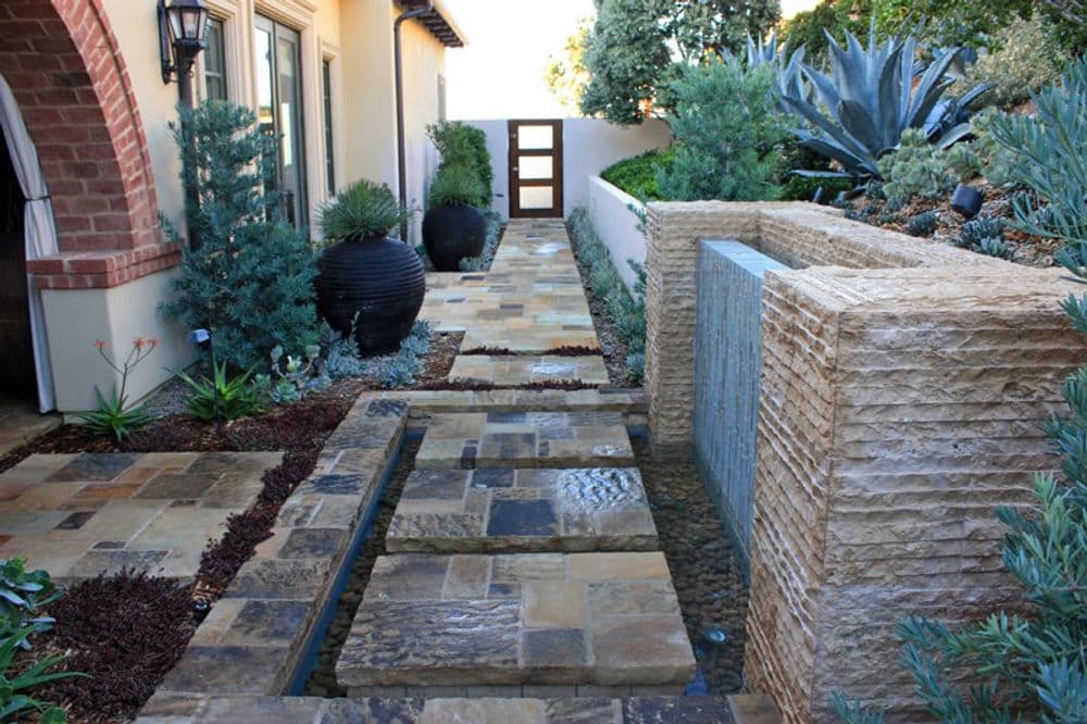 Modern garden pathway with stone tiles, greenery, and a sleek water feature.