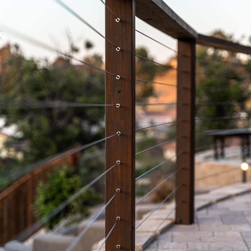 Wooden railing with metal cables along a landscaped pathway in a sunny outdoor setting.