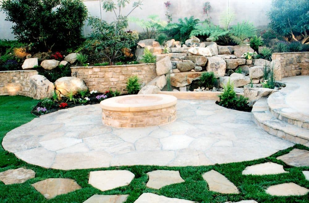 Elegant backyard landscape featuring a stone patio, fire pit, and natural rock waterfall.