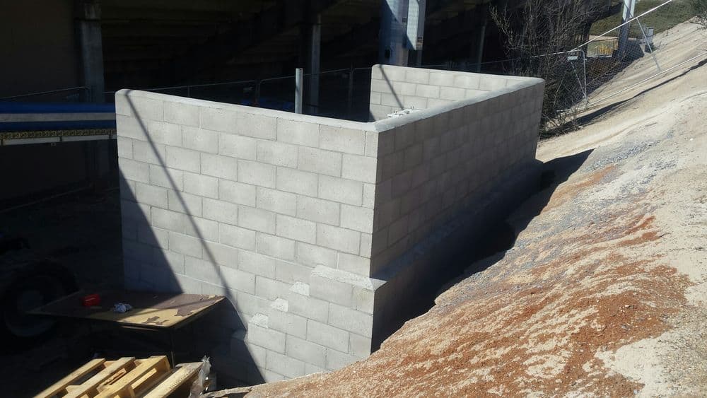 Cement block wall under construction near a sloped terrain and construction equipment.