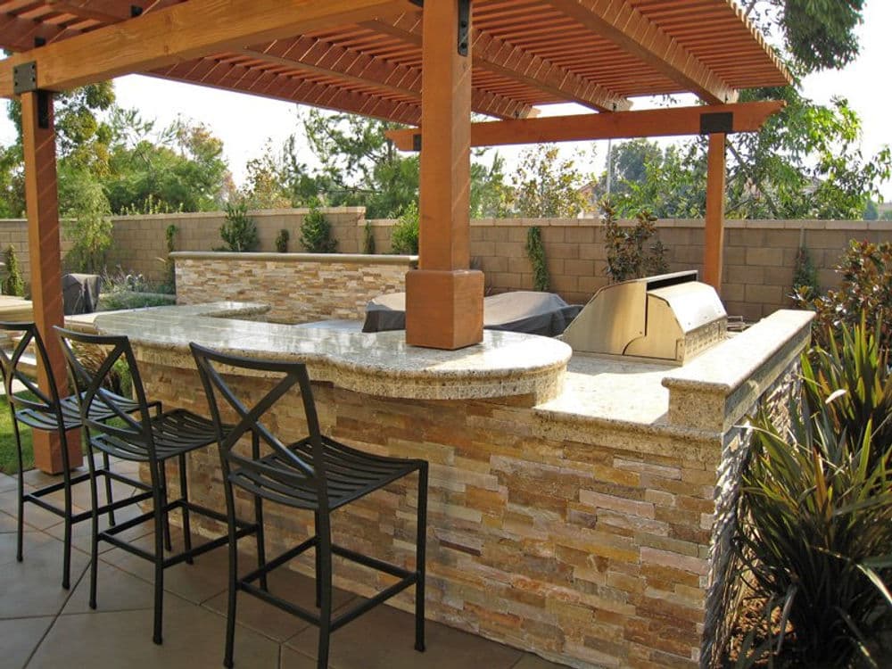 Outdoor kitchen with stone bar, grill, and seating beneath a wooden pergola. Perfect for gatherings.
