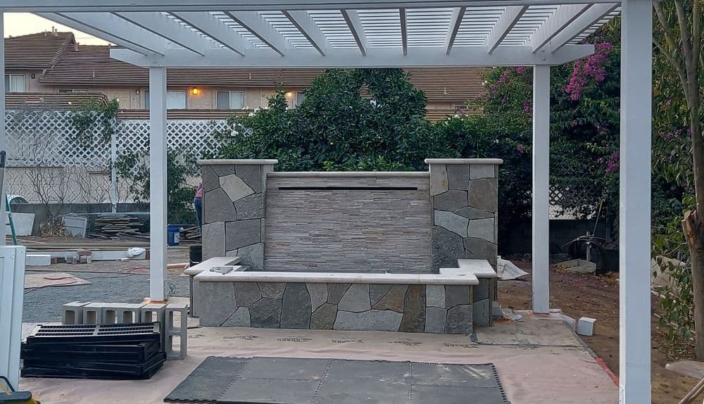 Outdoor stone fountain installation under a white pergola in a landscaped backyard.
