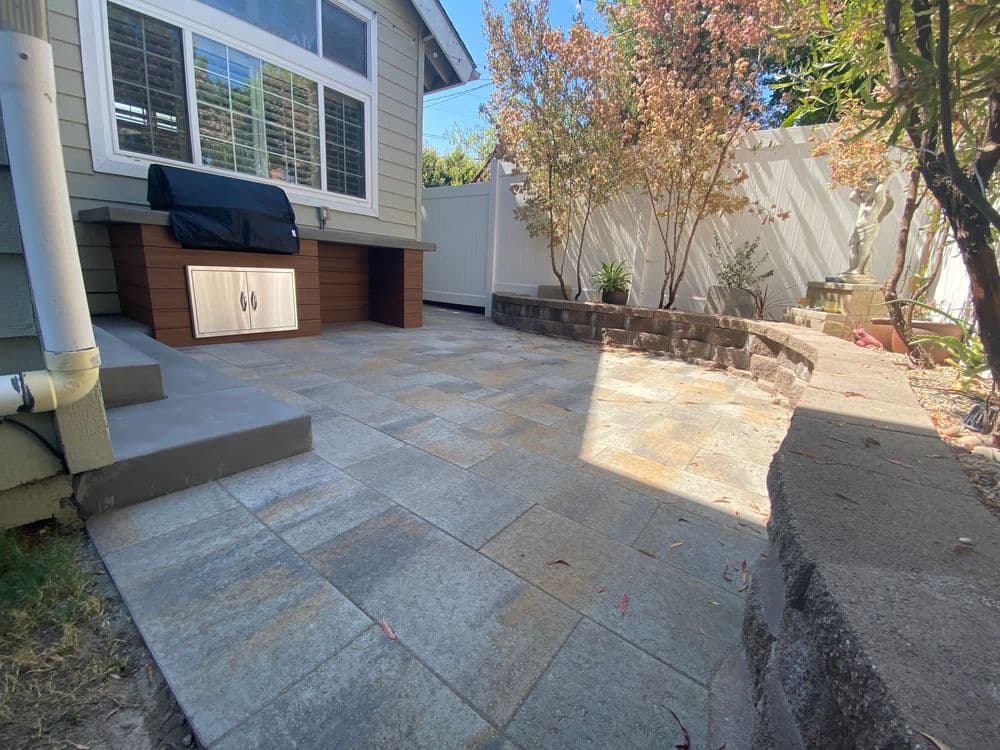 Patio area with stone tiles, seating, and plants surrounded by garden foliage and a fence.