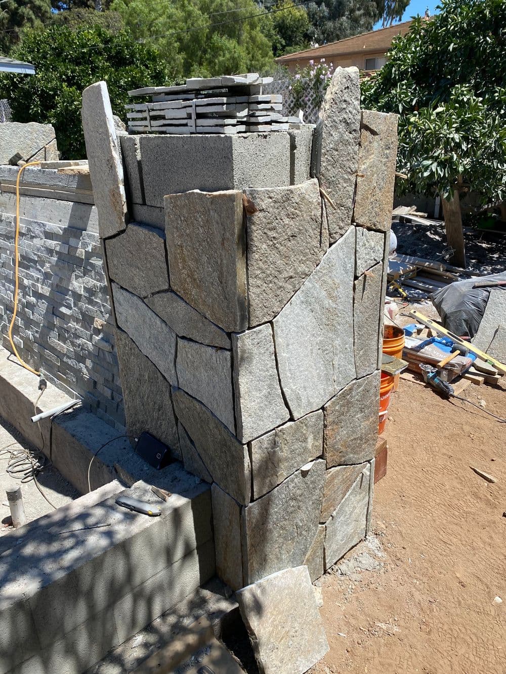 Natural stone wall with stacked stone blocks and construction materials in a backyard setting.