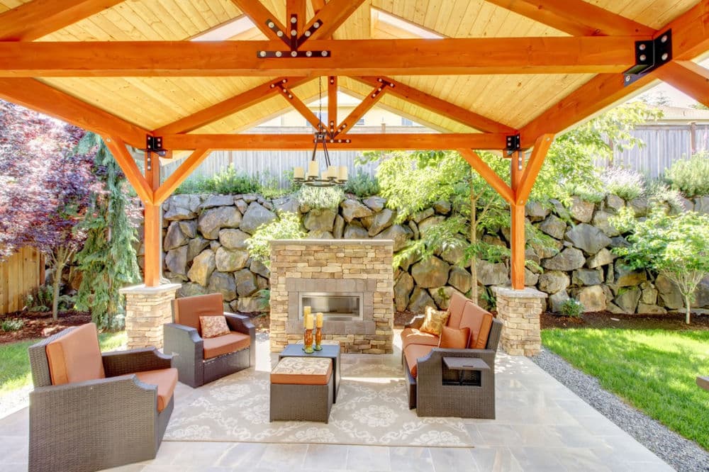 Outdoor patio with wooden pergola, stone fireplace, and cozy seating area surrounded by greenery.