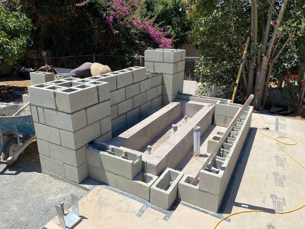 Cinder block wall construction with a stone feature and garden backdrop under clear blue skies.