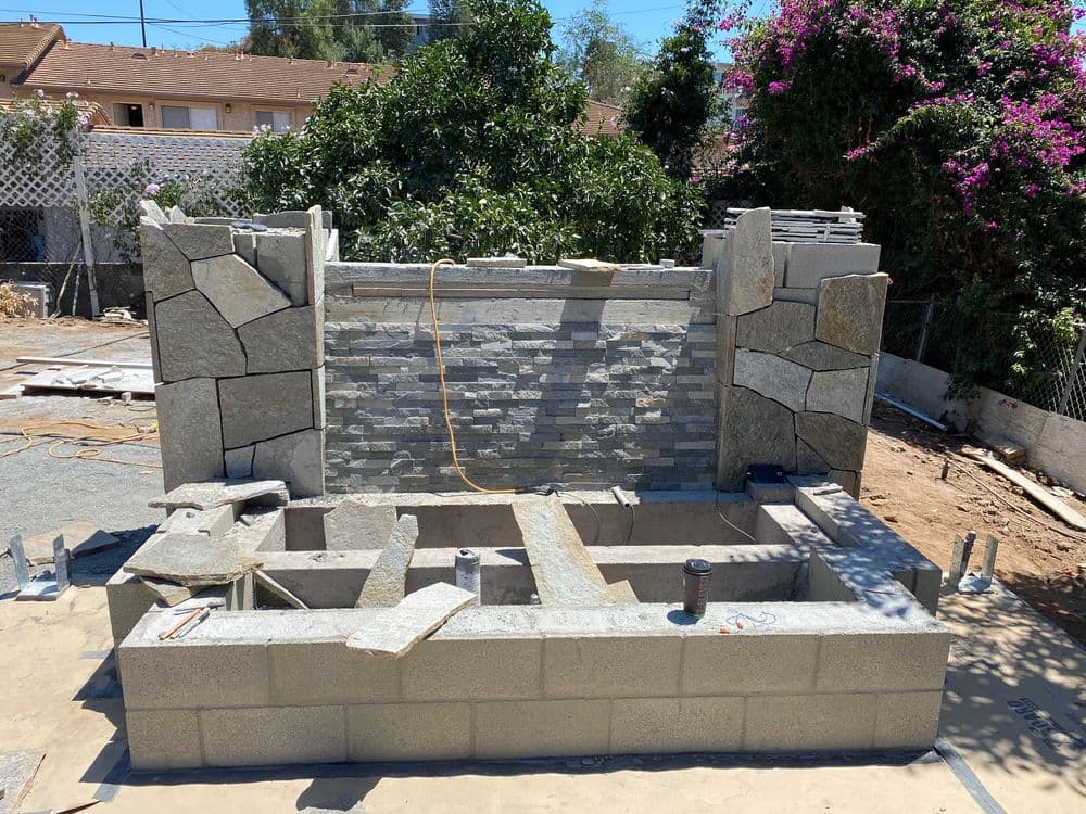 Stone outdoor fireplace construction with stacked stone and block layout in a garden setting.