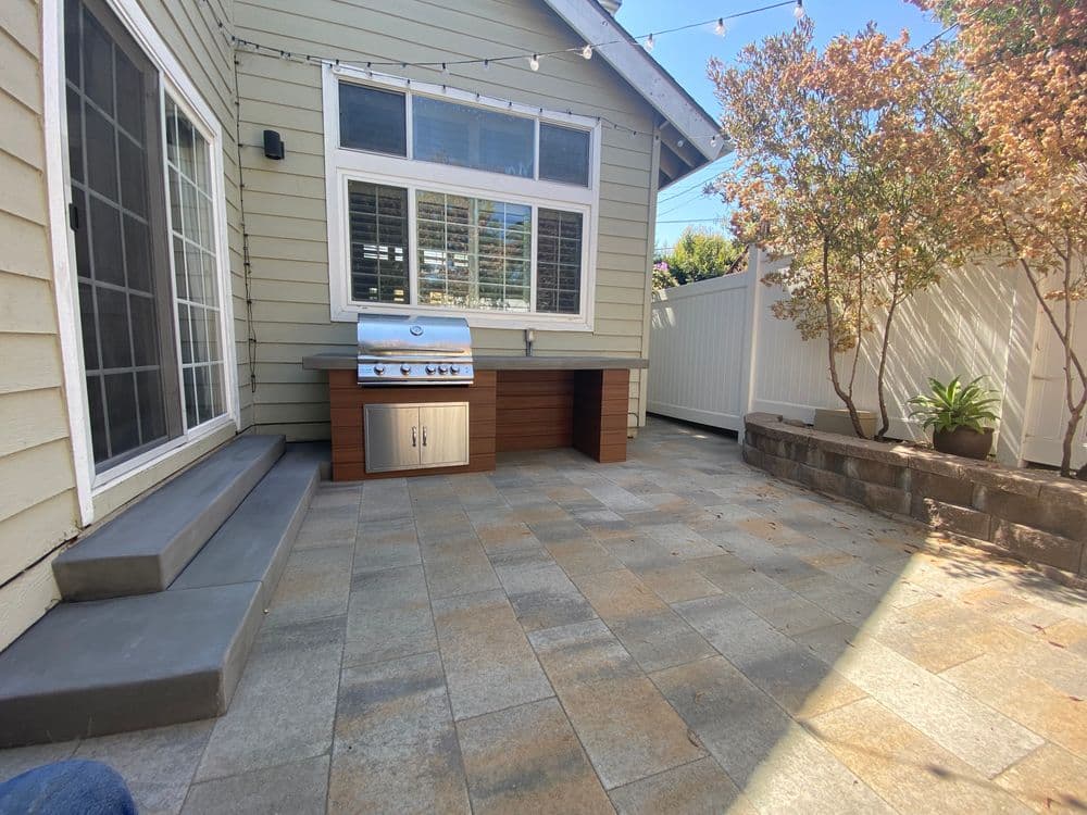 Backyard patio with a modern grill, stone pavers, and greenery in a sunny setting.