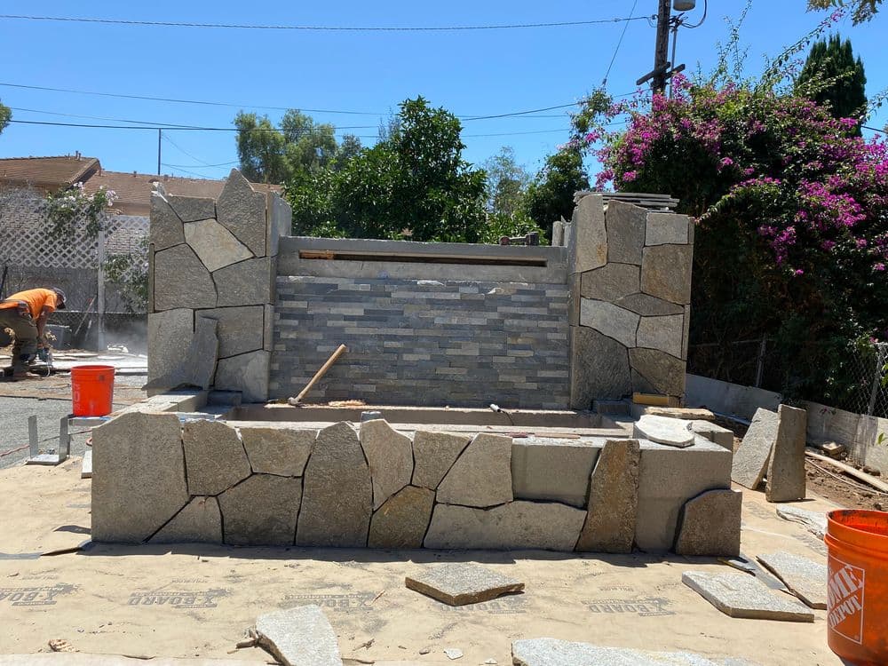 Stone wall installation with gray stones and patio area under construction in sunny outdoor setting.