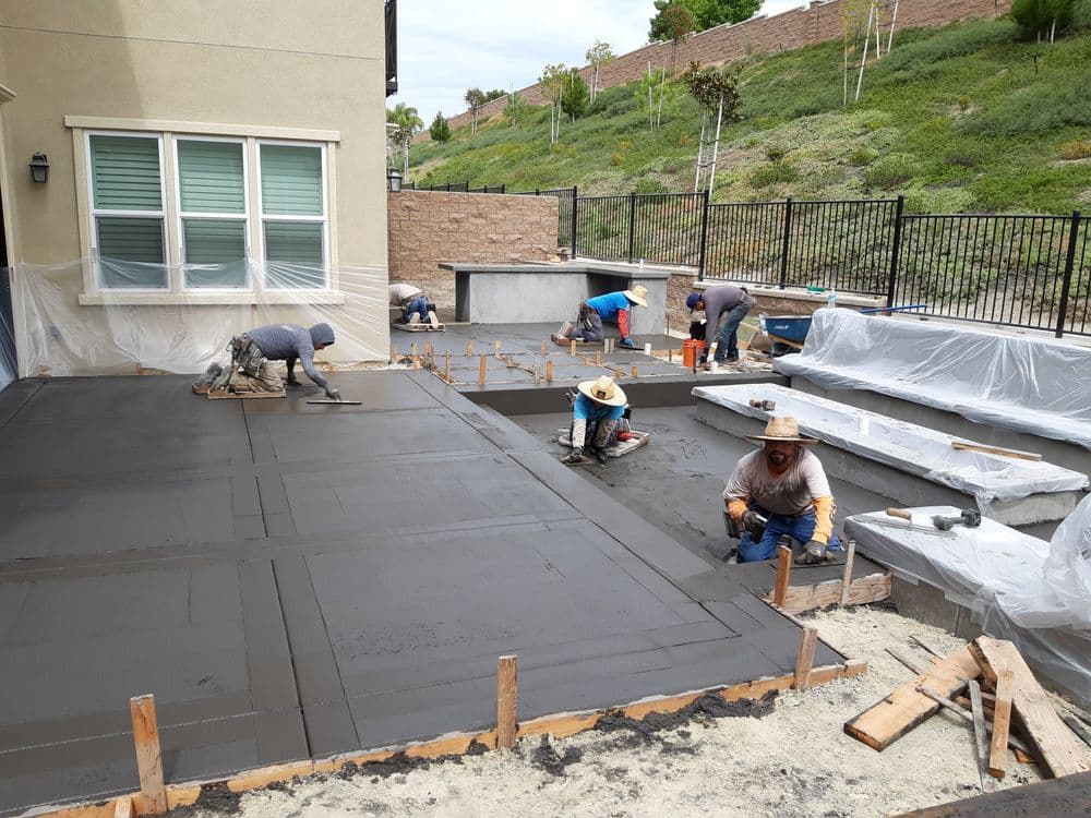Workers applying concrete in a backyard for patio construction with protective coverings.