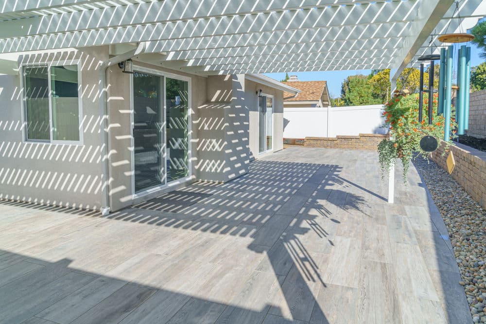 Bright patio with wooden pergola casting shadows, featuring stylish tile flooring and landscaping.
