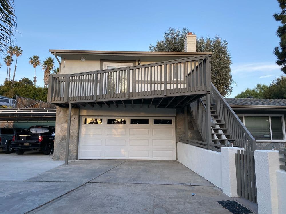 Two-story house with garage, stairs, and palm trees in the background under a clear sky.