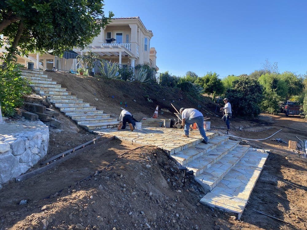 Workers installing stone steps in a residential landscaping project with a house in the background.