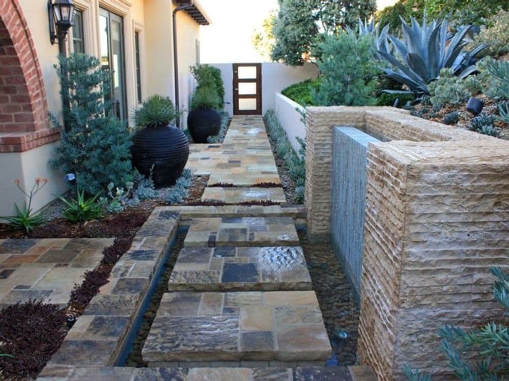 Modern landscaped pathway with stone tiles, water feature, and lush greenery.