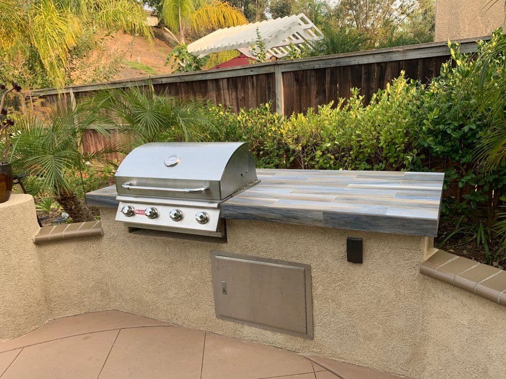 Outdoor kitchen with a stainless steel grill on a stone countertop, surrounded by greenery.
